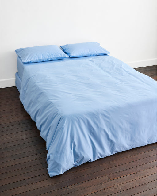 A neatly made bed features the Sky Organic Cotton Duvet Cover Set by Bed Threads in light blue, with two matching pillows on a dark wooden floor and a plain white wall background.