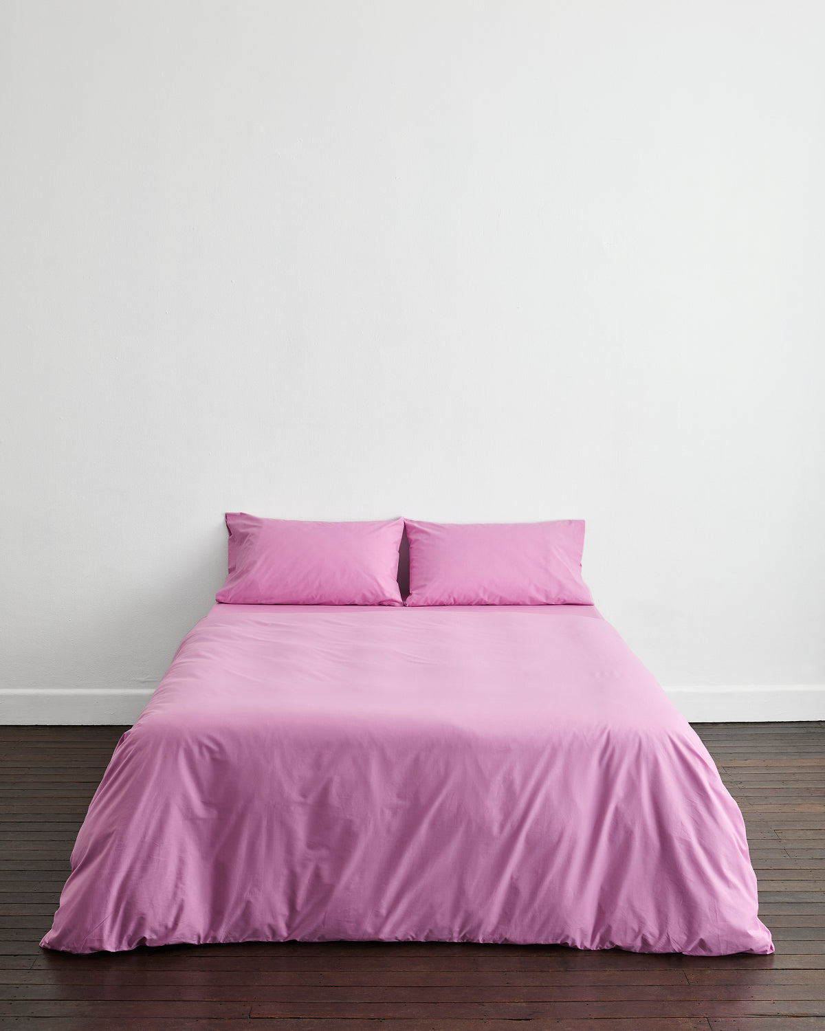 A neatly made bed features the Bed Threads Bubblegum Organic Cotton Bedding Set with two matching pillows, set against a plain white wall and wooden floor.