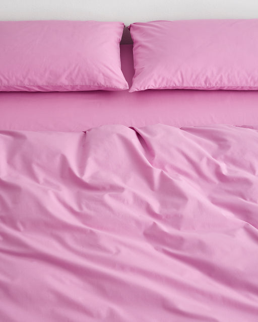 A neatly made bed with two pillows in Bed Threads Bubblegum Organic Cotton Pillowcases and a matching bright pink blanket, both slightly rumpled at the center. The pillowcases are crafted from breathable percale cotton.