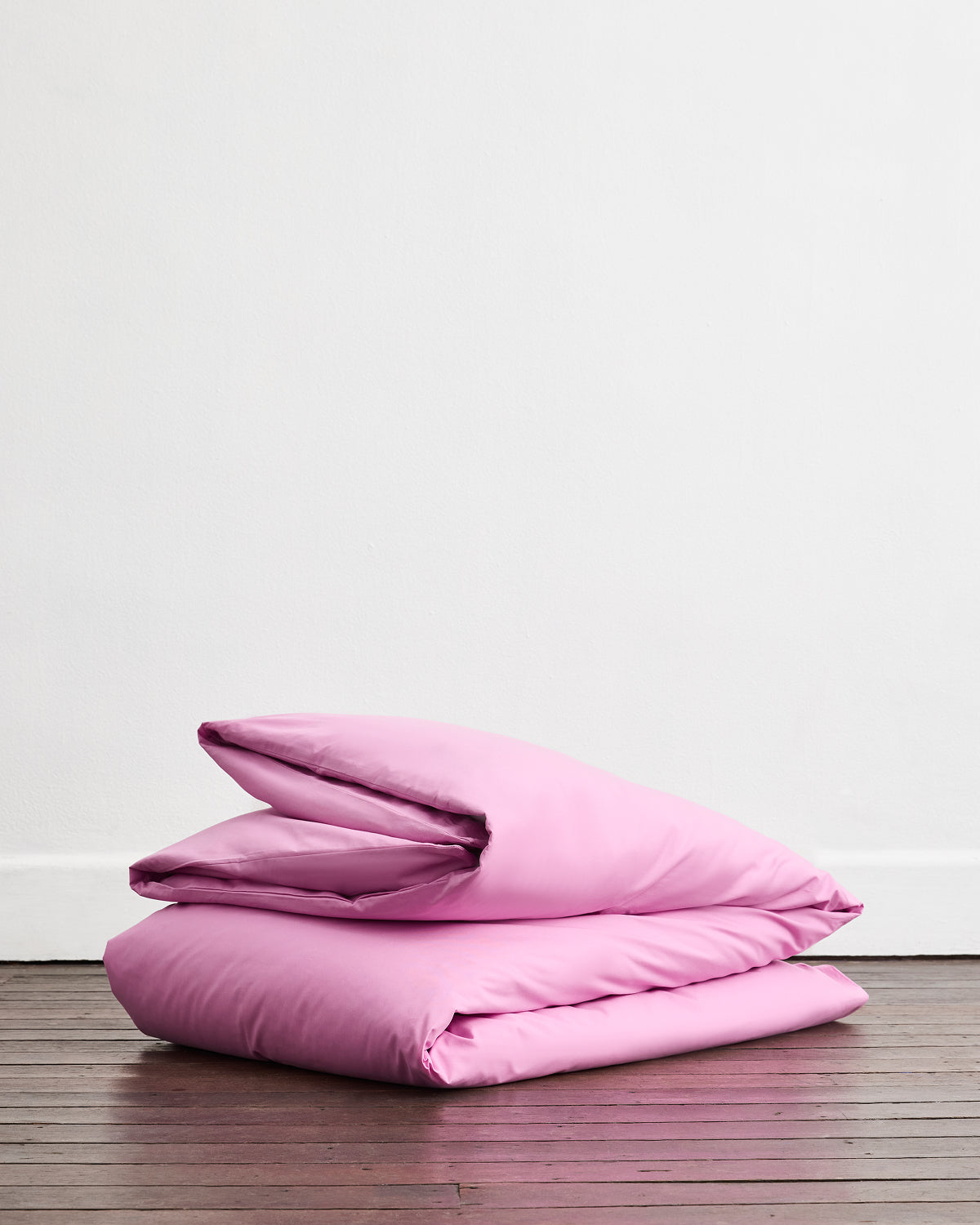 A Bubblegum Organic Cotton Duvet Cover by Bed Threads is neatly folded with a pillow on a wooden floor, set against a plain white wall.