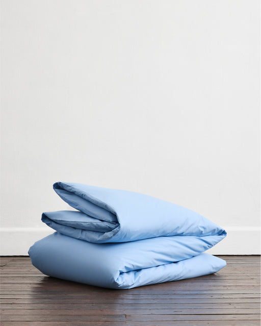 A neatly folded Sky Organic Cotton Duvet Cover Set by Bed Threads in light blue rests on a wooden floor against a plain white wall.