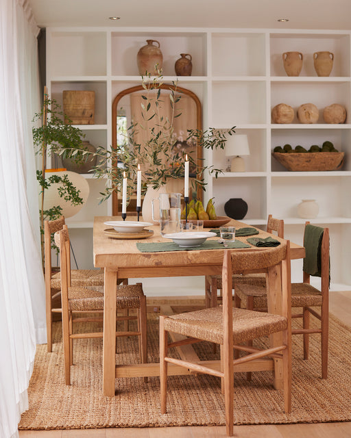 A wooden dining table set with plates, glasses, candles, and green napkins is surrounded by four woven chairs. Bed Threads Olive 100% French Flax Linen Placemats (Set of Four) enhance the rustic feel from pottery-filled shelves and lush greenery.