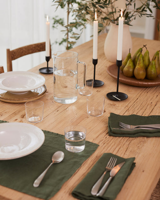 A wooden dining table set with white plates, green napkins, Bed Threads Olive 100% French Flax Linen Placemats (Set of Four), silverware, clear glasses, a glass pitcher of water, three lit candles, pears on a plate, and greenery in a vase.