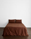 A neatly made bed with two pillows features the Bed Threads Espresso Organic Cotton Bedding Set, set against a plain white wall on a wooden floor.