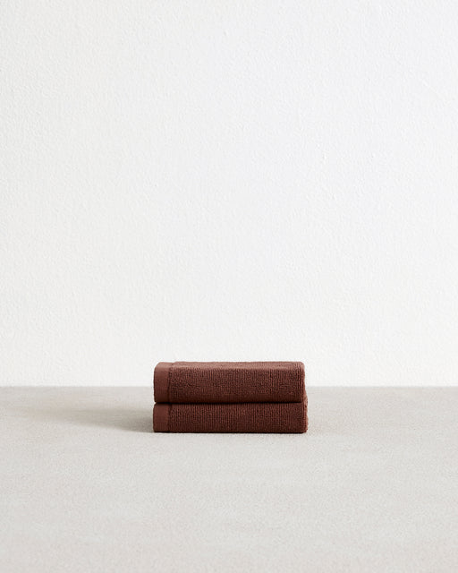 The Bed Threads Espresso 100% Cotton Terry Face Washers (Set of Two) feature dark brown rectangles with stitched edges and fabric labels, layered on a light surface. Made from absorbent terry, they're gentle on skin.