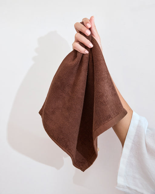 A folded Espresso 100% Cotton Terry Face Washer by Bed Threads sits on a white bathroom countertop beside a round sink and soap dish. The absorbent, gentle terry adds comfort and style to the modern, minimal setting. Set of two.