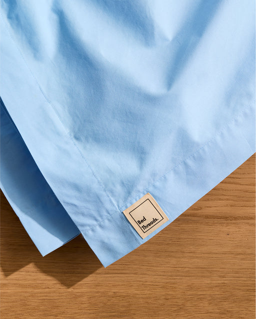 A Sky Organic Cotton Flat Sheet by Bed Threads, in light blue with a square beige "Red Threads" label, lies on a wooden surface.