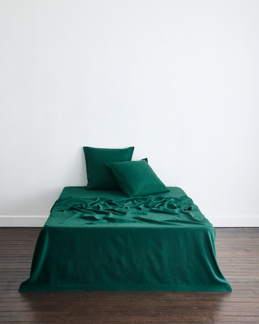 A neatly made bed with deep green sheets and two Forest 100% French Flax Linen European Pillowcases by Bed Threads, set against a white wall on a brown wooden floor. The bedding is slightly rumpled at the center.