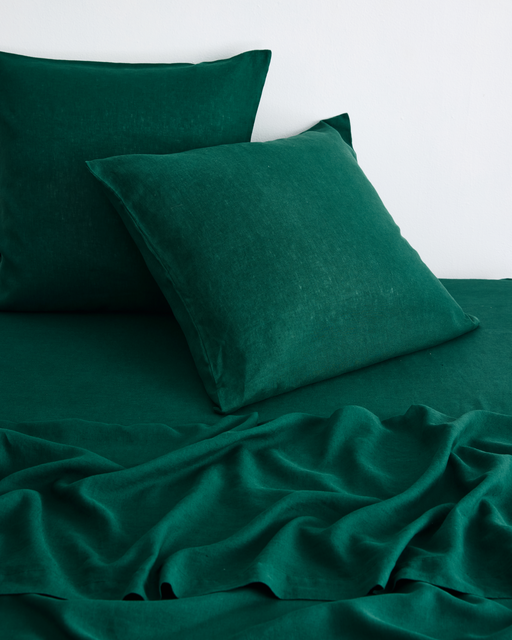 Two Bed Threads Forest 100% French Flax Linen European Pillowcases (Set of Two) in dark green rest on a matching linen sheet with a natural, slightly wrinkled texture against a white wall.