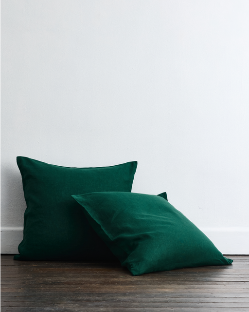 Two Forest 100% French Flax Linen European Pillowcases by Bed Threads rest against a white wall on a wooden floor, one upright and the other angled in front.