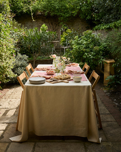 A rectangular outdoor table with a beige cloth is set for dining, featuring plates, bread, bowls, floral centerpieces, and Bed Threads Terracotta 100% French Flax Linen Napkins (Set of Four), surrounded by wooden chairs and greenery.