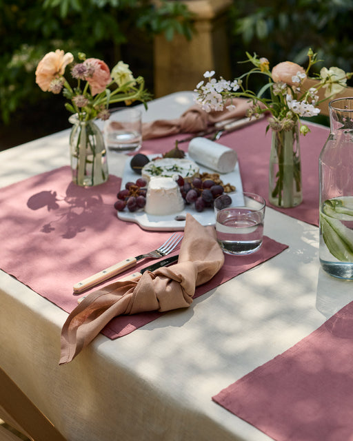 A sunlit outdoor table is set with blush pink placemats, Bed Threads’ Terracotta 100% French Flax Linen Napkins (Set of Four), glassware, fresh flowers in small vases, a carafe of cucumber water, and a cheese platter with grapes and nuts.