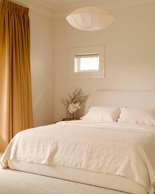 Minimalist bedroom with a light beige bed, Bed Threads’ Crème 100% French Flax Linen Flat Sheet, gold curtains, a small window, modern white pendant light, and a bedside table with a vase of white flowers. The palette is warm and neutral.
