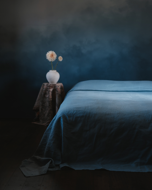 A neatly made bed with the Lagoon Heavy Linen Bed Cover by Bed Threads stands next to a round table draped in patterned fabric, topped with a white vase of light flowers, all set against a dark, moody blue background.