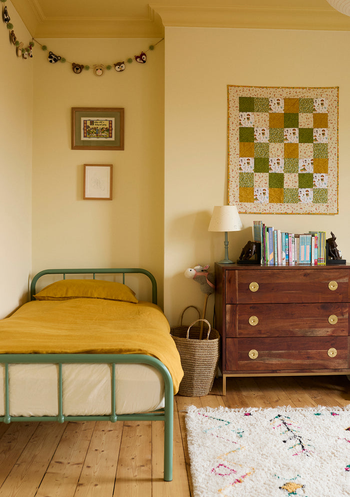 A cozy bedroom with a green metal bed frame, mustard yellow bedding, a wooden dresser with books and a lamp, wall art, a garland, and a woven basket, all on a light wood floor with a white patterned rug.