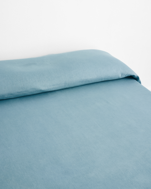 A neatly made bed features the Bed Threads Lagoon Heavy Linen Bed Cover in blue, paired with a matching pillow against a white wall, highlighting the soft elegance of heavyweight linen bedding.