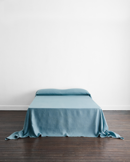 A minimal bed features the Bed Threads Lagoon Heavy Linen Bed Cover in light blue, set against a white wall on a dark wooden floor, with the cover draped loosely at the corners.