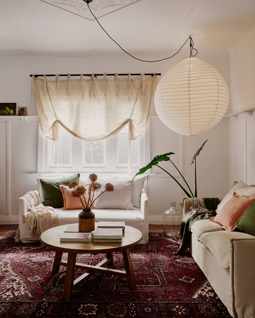 A cozy living room with two sofas topped with Bed Threads’ Olive 100% French Flax Linen European Pillowcases (Set of Two), a round wooden coffee table, dried flowers in a vase, a large paper lantern ceiling light, and daylight through beige curtains.