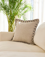 A Bed Threads Oatmeal & Charcoal 100% French Flax Linen Swirl Cushion Cover with decorative dark trim rests on a cream sofa, with sunlight shining through sheer curtains and a tall green plant in the background. - view