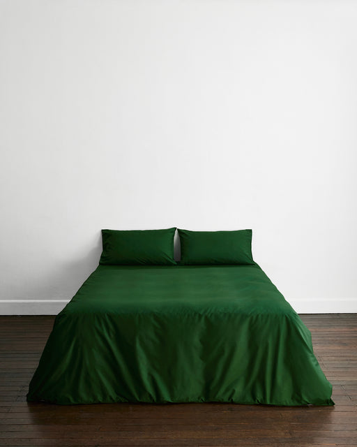 A minimalist room features a neatly made bed with dark green bedding and two Bed Threads Moss Organic Cotton Pillowcases, centered against a plain white wall on a wooden floor.