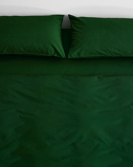 A bed with Bed Threads’ Moss Organic Cotton Pillowcases (Set of Two), dark green sheets, a green duvet, and a plain white wall backdrop.