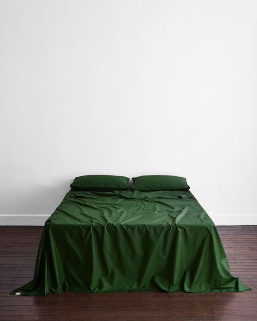 A neatly made bed features the Moss Organic Cotton Flat Sheet and two matching pillows by Bed Threads, centered against a plain white wall on dark wooden floors—ideal for anyone who values breathable organic bedding.