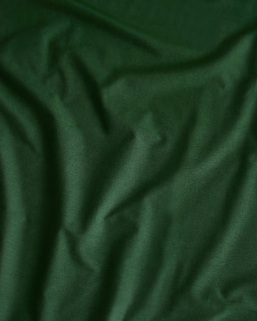 A close-up of the Moss Organic Cotton Flat Sheet by Bed Threads shows its dark green percale fabric with visible creases, highlighting the breathable, textured quality of this organic bedding.