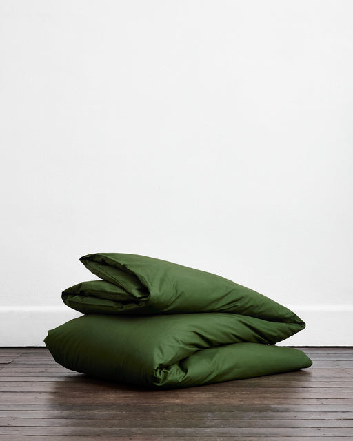 Two Moss, Apple & Crisp White Organic Cotton Bedding Bundles by Bed Threads are neatly folded and stacked on a wooden floor against a plain white wall.