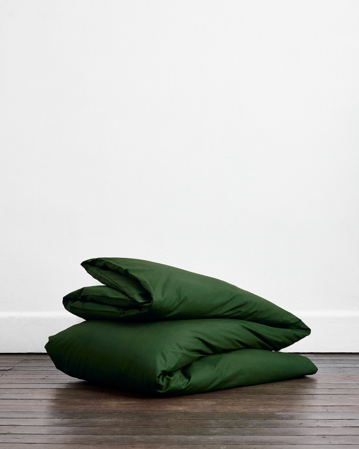 Two dark green pillows rest on a wooden floor against a white wall, perfectly matching the cozy vibe of the Bed Threads Moss Organic Cotton Duvet Cover Set.