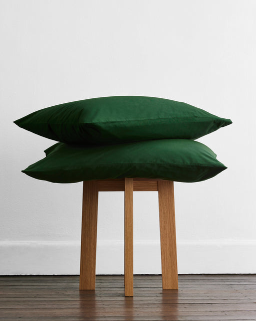 Two dark green pillows rest on a wooden stool, emphasizing the cozy charm of the Bed Threads Apple & Moss Organic Cotton Bedding Bundle, with a plain white wall and dark wood floor as a backdrop.