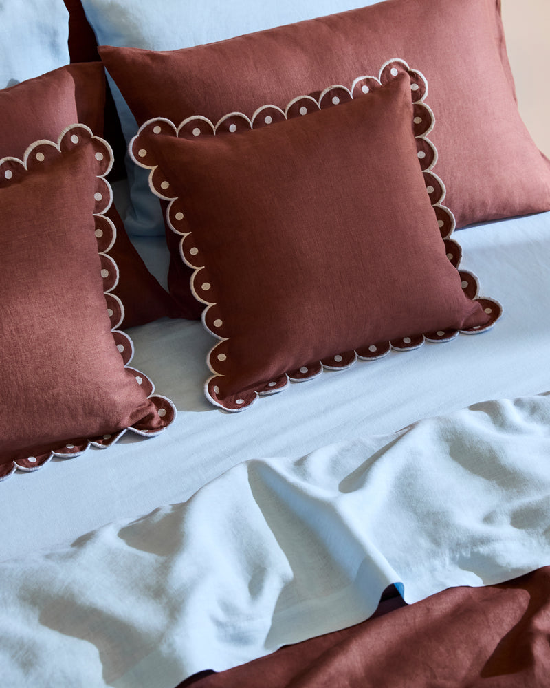 Two maroon decorative pillows with white scalloped edges and a Cacao & Coast 100% French Flax Linen Arch Cushion Cover by Bed Threads rest on light blue sheets and maroon pillowcases, creating a cozy, contrasting look.