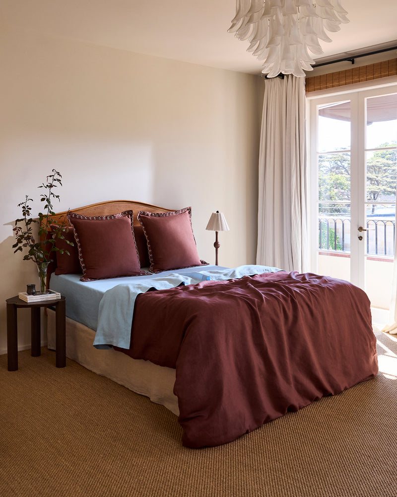 A neatly made bed features Bed Threads’ Cacao & Coast 100% French Flax Linen Swirl European Pillowcases (Set of Two) with decorative edging, a light blue linen sheet, a brown blanket, and a small lamp on the wooden nightstand.