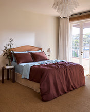 A neatly made bed with light blue sheets and Bed Threads’ Cacao & Coast 100% French Flax Linen Swirl Pillowcases, featuring brown embroidery and white loops, sits against a wooden headboard. The linen bedspread has a brown section at the bottom. - view