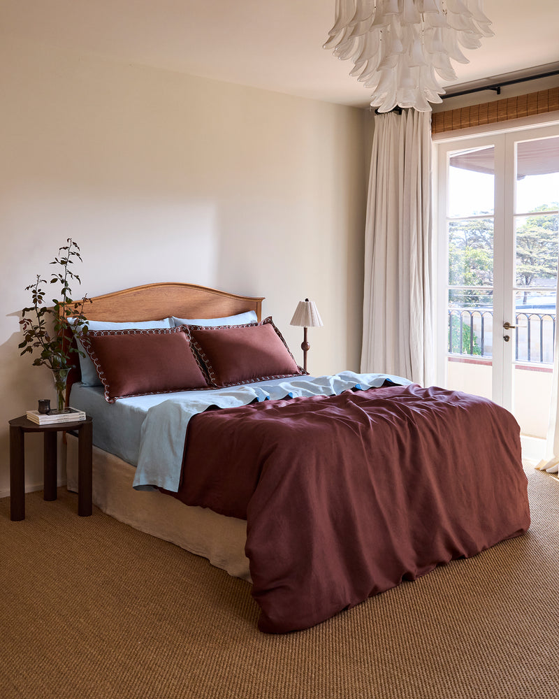 A neatly made bed with light blue sheets and Bed Threads’ Cacao & Coast 100% French Flax Linen Swirl Pillowcases, featuring brown embroidery and white loops, sits against a wooden headboard. The linen bedspread has a brown section at the bottom.