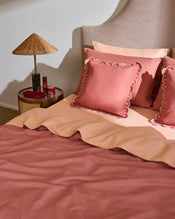 A neatly made bed with rose and blush bedding, scalloped-edge pillows, and a headboard is accented by the Bed Threads Pink Clay & Terracotta 100% French Flax Linen Arch Cushion Cover. A round side table holds a wicker lamp, candle, and red bottle for a cozy touch. - view