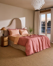 A neatly made bed features Bed Threads’ Terracotta & Pink Clay 100% French Flax Linen Swirl Pillowcases (set of two) with embroidered edges, a matching peach bedspread, and a reddish-pink linen sheet, all set against a beige headboard. - view