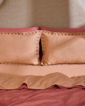 A neatly made bed features Bed Threads’ Terracotta & Pink Clay 100% French Flax Linen Swirl Pillowcases (set of two) with embroidered edges, a matching peach bedspread, and a reddish-pink linen sheet, all set against a beige headboard. - view