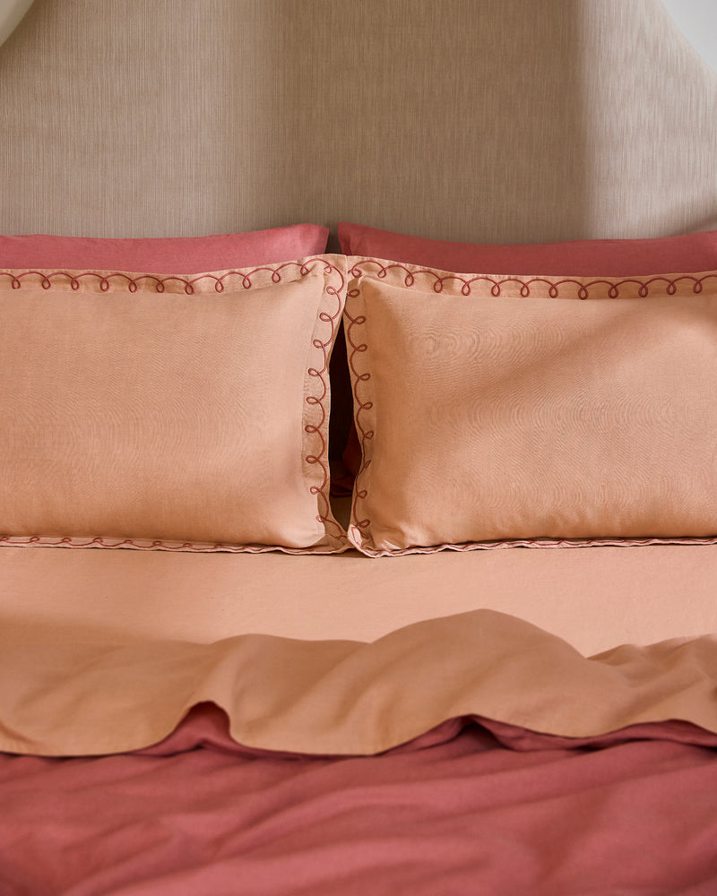 A neatly made bed features Bed Threads’ Terracotta & Pink Clay 100% French Flax Linen Swirl Pillowcases (set of two) with embroidered edges, a matching peach bedspread, and a reddish-pink linen sheet, all set against a beige headboard.