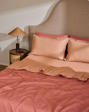 A neatly made bed features Bed Threads’ Terracotta & Pink Clay 100% French Flax Linen Swirl Pillowcases (Set of Two), arranged with two pillows, a curved beige headboard, and a round wooden nightstand with a woven-shade lamp. - view