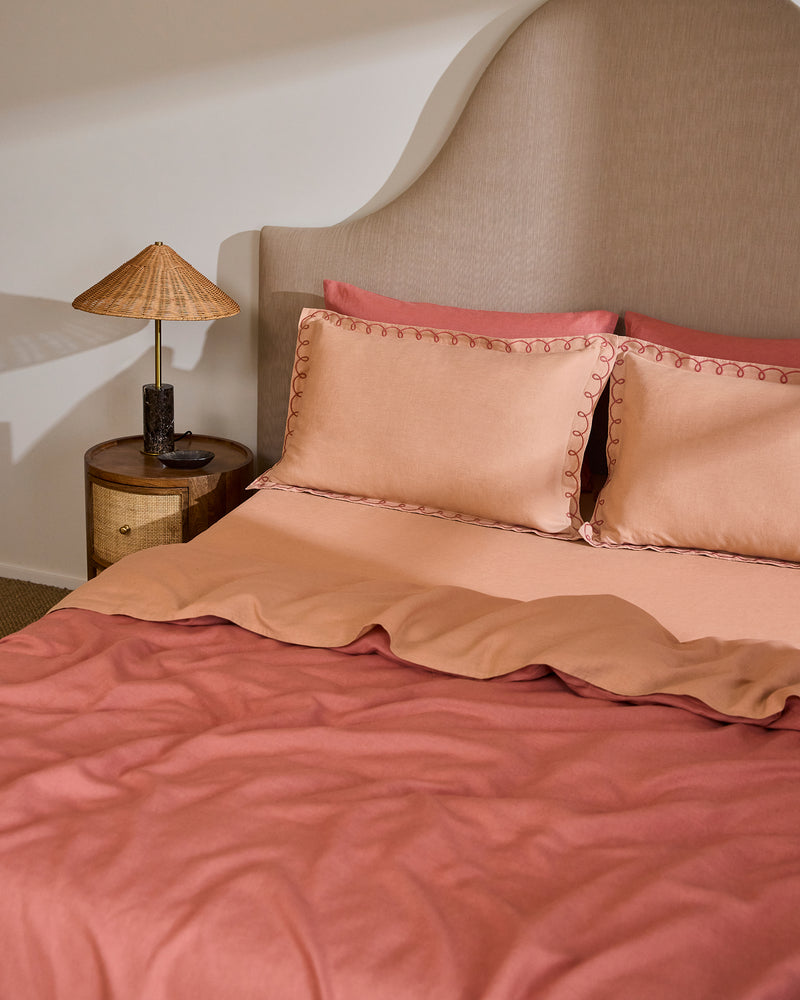 A neatly made bed features Bed Threads’ Terracotta & Pink Clay 100% French Flax Linen Swirl Pillowcases (Set of Two), arranged with two pillows, a curved beige headboard, and a round wooden nightstand with a woven-shade lamp.