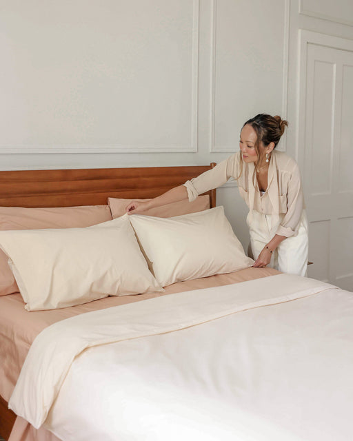 A woman in light clothing is making a neatly arranged bed with cream sheets and Bed Threads' Vanilla Organic Cotton Pillowcases (Set of Two) in a bright, minimal bedroom.