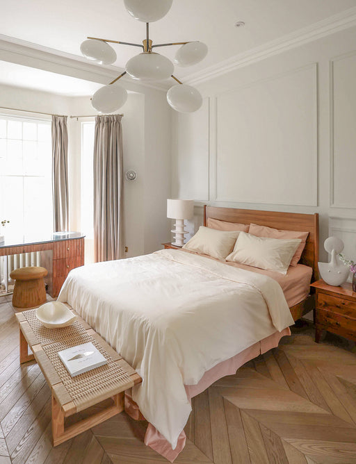 A bright, modern bedroom features a wooden bed dressed in Bed Threads’ Butterscotch Organic Cotton Fitted Sheet, Vanilla bedding, a wooden bench at the foot, two nightstands, gray curtains on a large window, and a contemporary white ceiling light.