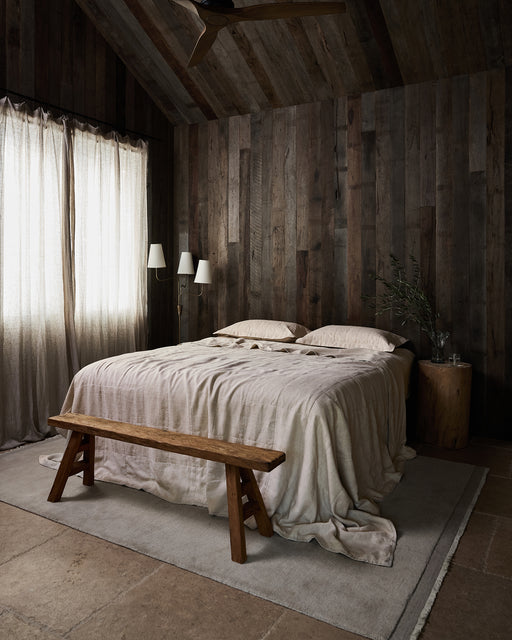 A rustic bedroom with dark wood-paneled walls and ceiling, a bed dressed in the Oatmeal Linen Quilt by Bed Threads, a wooden bench at the foot, tree-stump nightstand, tall curtains, floor lamp, and neutral rug on stone flooring.