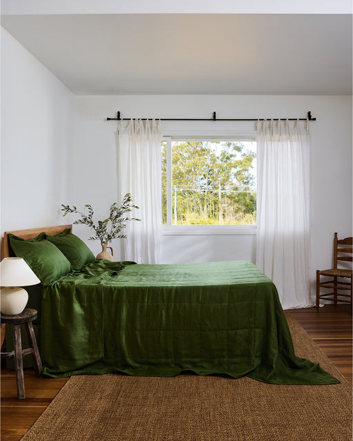 Bright bedroom with a large window, sheer white curtains, a bed dressed in the Olive Quilt by Bed Threads, a small wooden stool and lamp, wooden chair, and woven rug on the floor. Greenery is visible outside.