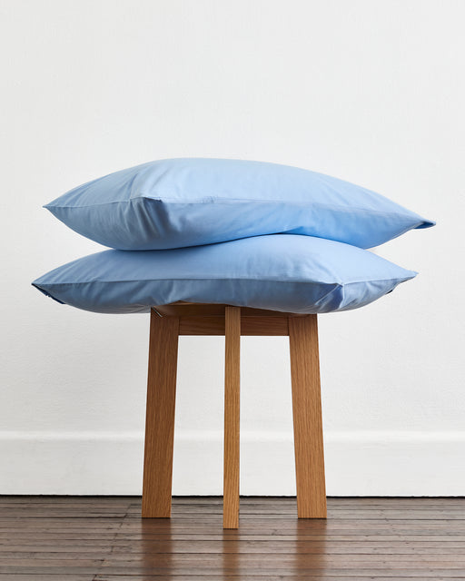 Two light blue pillows from the Bed Threads Espresso, Sky & Crisp White Organic Cotton Bedding Bundle are stacked on a four-legged wooden stool against a white wall and wooden floor.