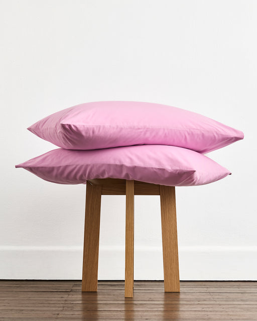 A pair of Bubblegum Organic Cotton Fitted Sheet Sets from Bed Threads is stacked on a small wooden stool, set against a white wall and wooden floor.