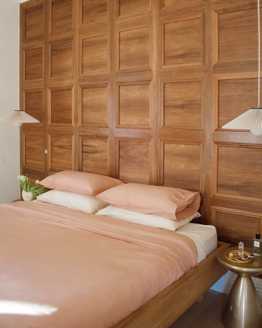 A cozy bedroom with a wood-paneled accent wall, a bed made up with the Vanilla Organic Cotton Flat Sheet by Bed Threads in peach, two pendant lights, a gold round nightstand, and a vase of white tulips.