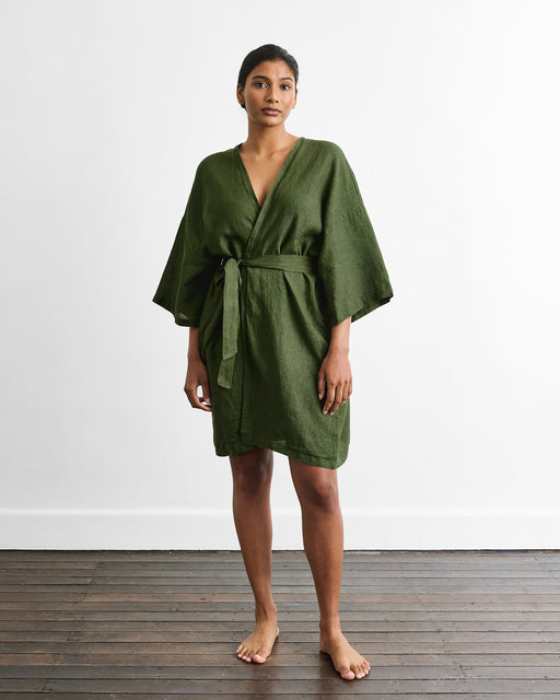 A person stands barefoot on a wooden floor against a plain white wall, wearing the Bed Threads Cabana Short Robe in Olive—a loose-fitting, knee-length linen robe with a belt.