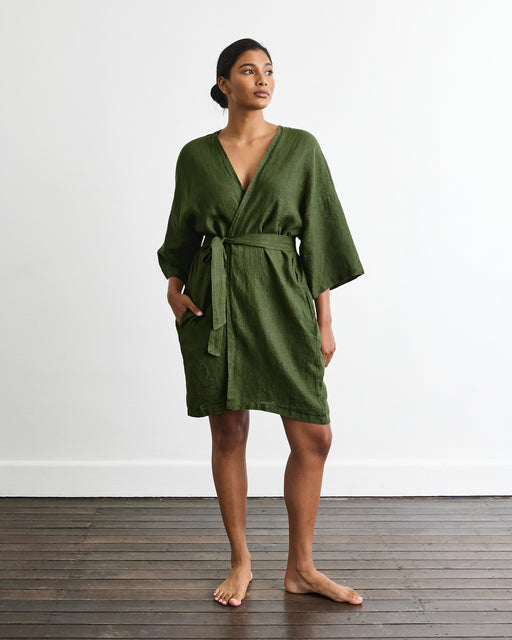 A woman stands barefoot on a wooden floor by a white wall, wearing the Bed Threads Cabana Short Robe in Olive—a loose, knee-length linen robe with a belt. Her hands rest in the pockets as she gazes slightly to the side.