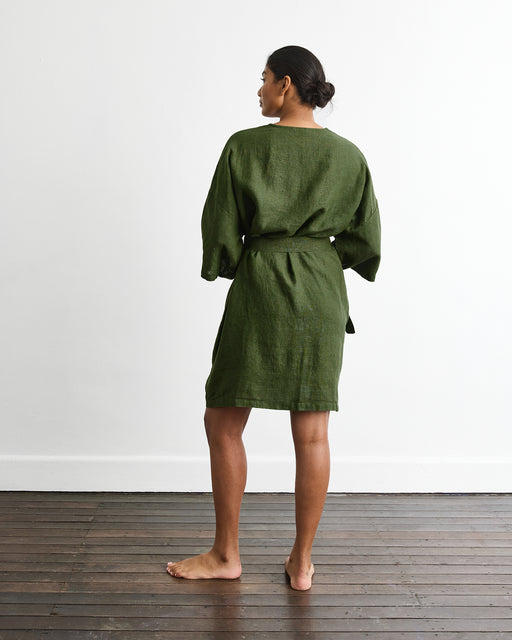 A person stands barefoot on a wooden floor, facing away, wearing the Bed Threads Cabana Short Robe in Olive with three-quarter sleeves. The background is a plain white wall.